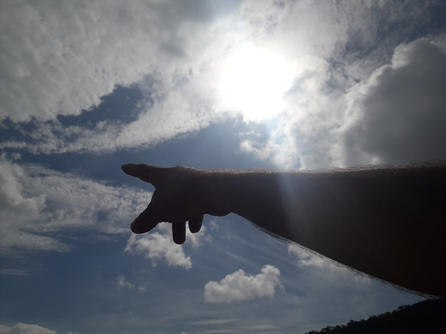Brazo señalando al cielo con nubes y el sol brillando al fondo